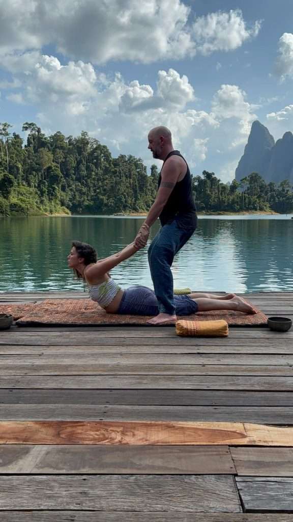 On a peaceful lakeside jetty in Wales, a caring guide gently supports a woman's relaxing Mountain Flow Massage-inspired stretch.