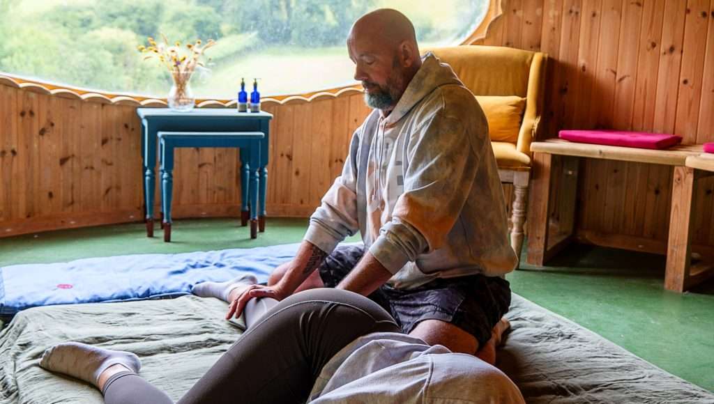 A caring bearded therapist gently works on a client’s lower back in a warm, peaceful room with cosy Welsh-inspired touches.